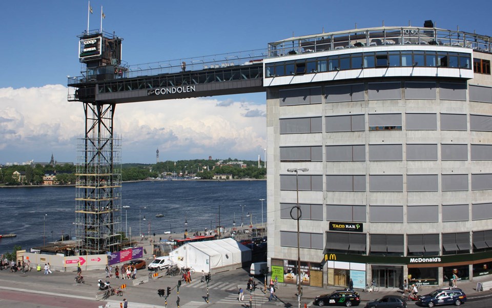 Katarinahissen sedd från Söder. Runt hissens ståltorn är byggställningar monterade och runt botten är det byggplank. Runt om området rör sig människor och trafik.